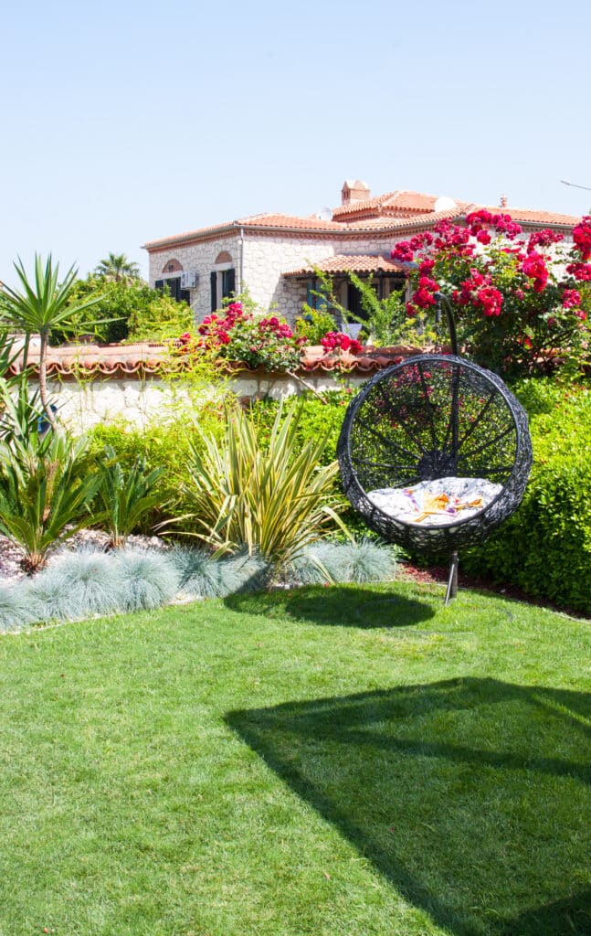 Jardin fleuri avec chaise suspendue et maison traditionnelle Jardin luxuriant avec chaise suspendue en osier noir, haies vertes, fleurs rouges et maison en pierre à toit de tuiles.