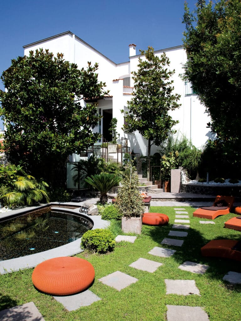 Jardin moderne avec bassin et mobilier orange vif Jardin luxuriant avec bassin sombre, pelouse, mobilier extérieur orange et maison blanche sous un ciel bleu.