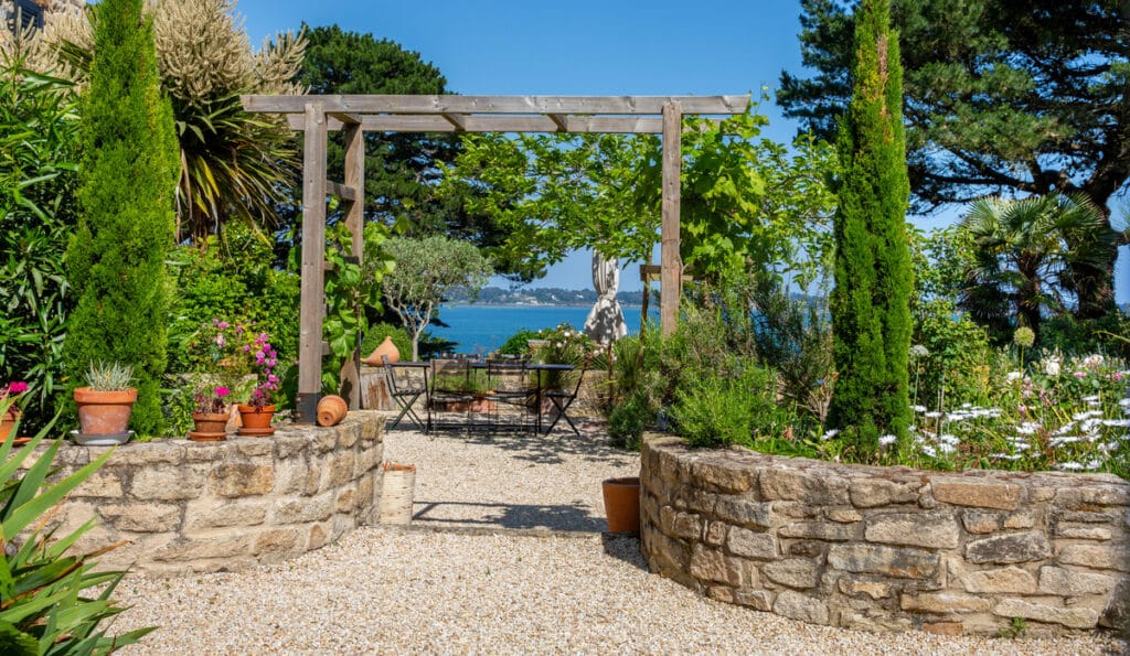 Jardin avec vue mer, pergola et mobilier extérieur Terrasse en gravier avec pergola en bois, muret en pierre, table et chaises offrant une vue sur une mer bleue et des jardins luxuriants.