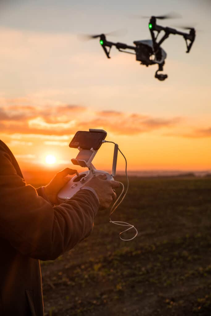 Pilotage Drone au Coucher de Soleil sur Champ Un pilote contrôle un drone en vol au coucher du soleil orange avec les mains sur la télécommande connectée à un smartphone.