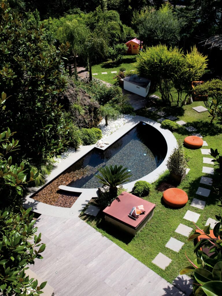 Jardin piscine contemporain : détente et verdure luxuriante Vue plongeante sur une piscine moderne aux mosaïques sombres, entourée de verdure luxuriante, d'une terrasse en dalles grises et de poufs orange.